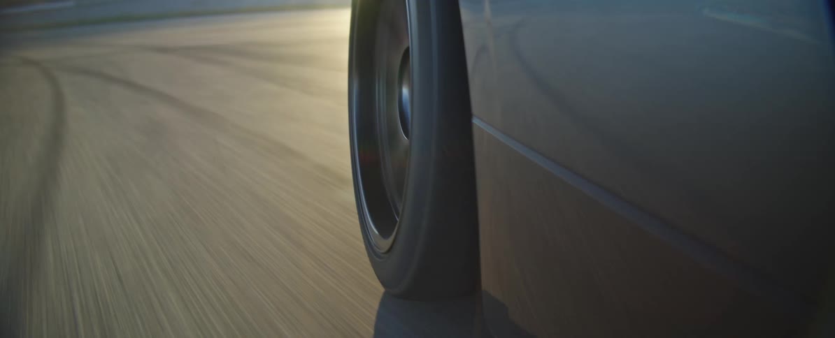 A closeup shot captures the spinning rear wheel of a dark car as it speeds along a curved asphalt track sunlight glints off the vehicles body and creates lens flares as the car navigates the bend at high speed