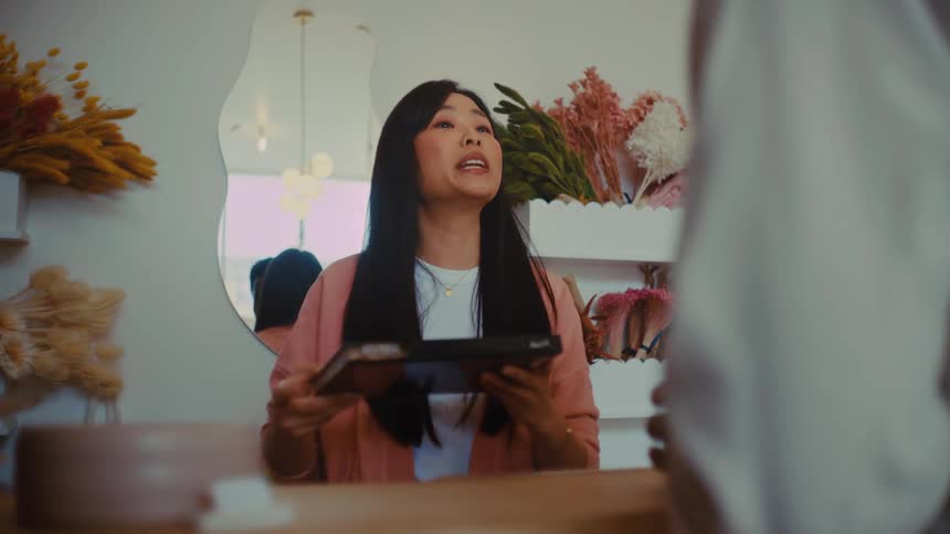 A cashier stands at the counter at midday handing a notebook to a customer with a warm smile they share a brief friendly exchange as the light from the storefront windows brightens the space around them