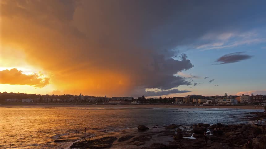People move on the shore as storm clouds flow over the nearby town covering the sun sitting on the horizon