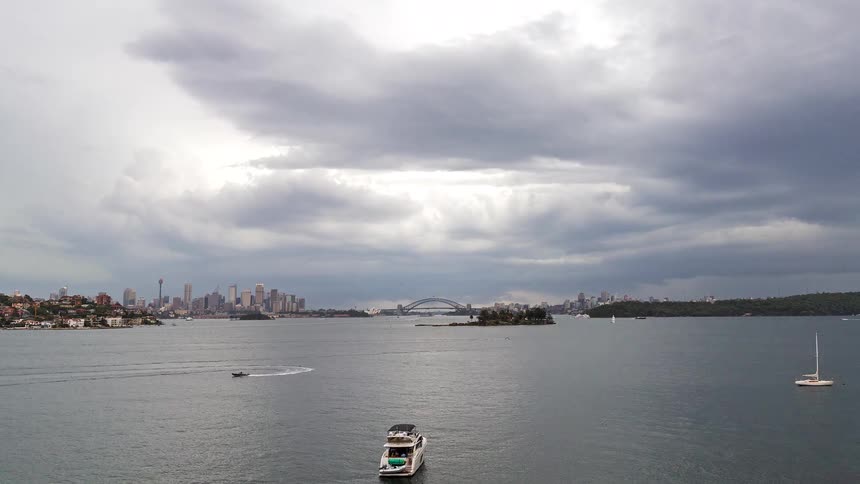 A distant view of the sydney city skyline and harbor bridge under a dark stormy sky as rain begins to fall and a white yacht floats on the water in the foreground
