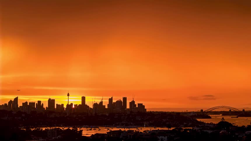 A city skyline filled with rows of skyscrapers and highrise buildings stretches toward the ocean the water visible in the distance as time moves from midday to sunset and later night settles in as the buildings glow
