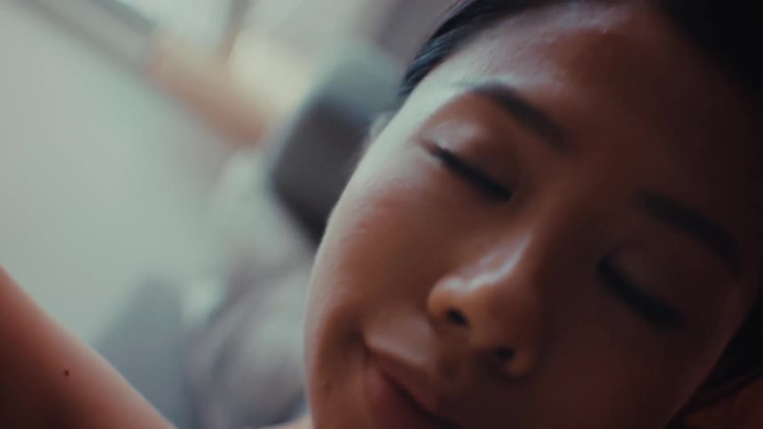 A close view of a young woman with her eyes closed as she sits in a room and smiles while tilting her head