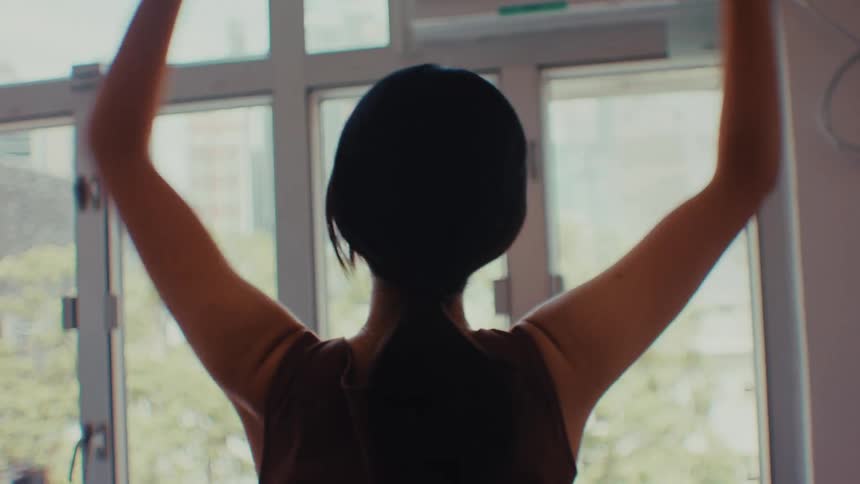 Woman performs arm stretches with arms raised near a window in a welllit environment