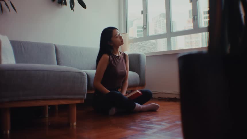 A woman wearing a brown sleeveless top and black pants sits on a wooden floor in a living room as she closes a red book and begins to stretch her legs and torso by a gray sofa