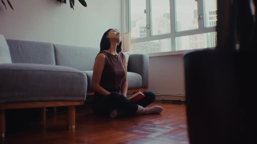 A woman wearing a brown sleeveless top and black pants sits on a wooden floor in a living room as she closes a red book and begins to stretch her legs and torso by a gray sofa