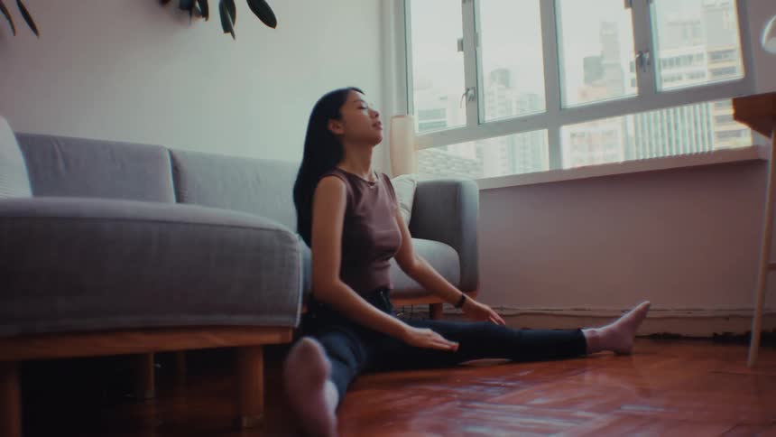 A young woman sits on a wooden floor in a sunlit living room performing various stretching exercises