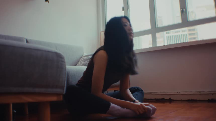 A young woman sits on a wooden floor in a sunlit living room performing various stretching exercises