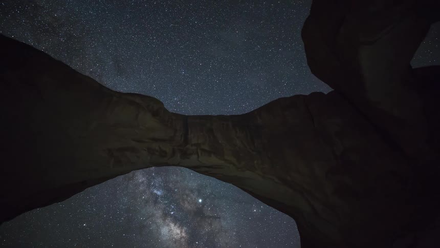 A view from below a large natural stone arch looks up at a dark night sky filled with a vast number of stars and the glowing milky way galaxy