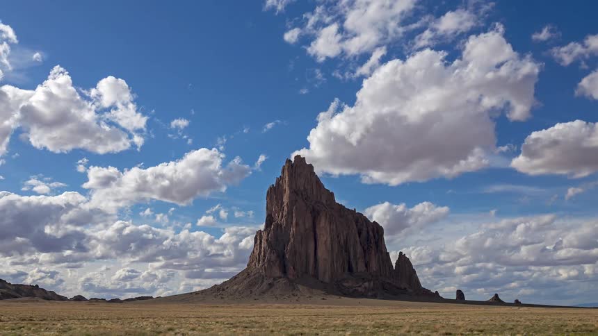 A distant view shows a large jagged rock peak standing in a flat desert plain under a blue sky with many white clouds moving quickly across the landscape