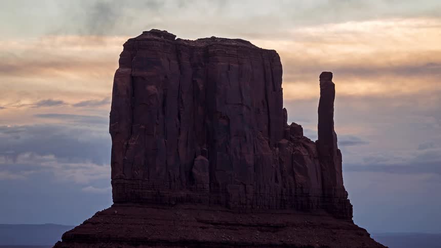 A distant view of a large dark rock butte stands against a cloudy sky with orange and blue tones at dusk in a desert landscape