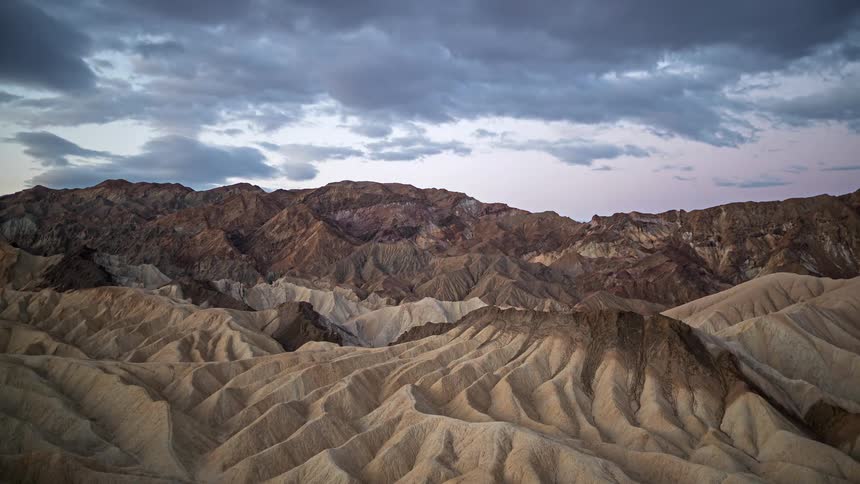 Undulating desert formations extend toward a rocky mountain range under a cloudy sky