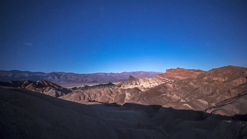 A distant view of a desert mountain range with sharp ridges and dark shadows under a clear blue night sky filled with stars