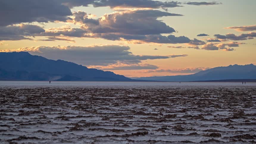A distant view of a vast salt flat with a textured surface under a sunset sky with golden clouds and dark mountain silhouettes in the background