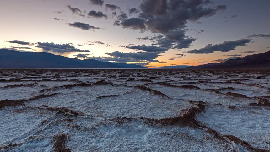 Snow covered rough terrain with mountains in the distance as clouds move overhead
