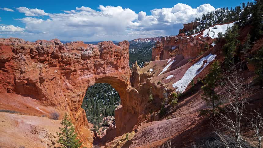 Natural red rock arch stands out in a canyon with vegetation and patches of snow