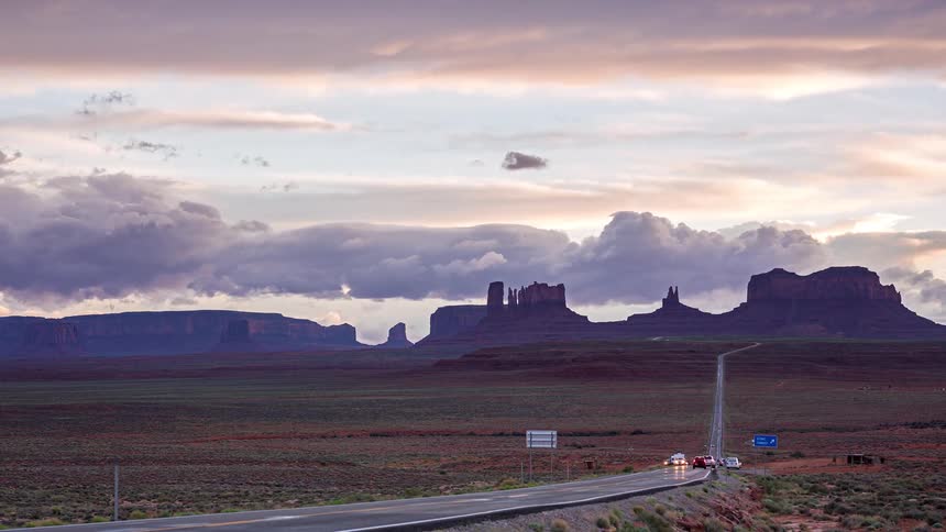 Rock formations rise in the background as a straight road crosses monument valley under a cloudy sky