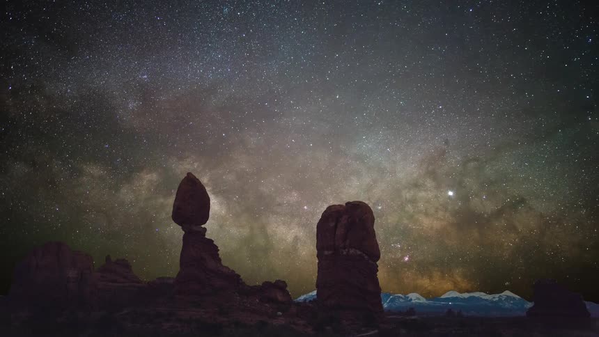 A view of a night sky full of stars above a desert landscape with large rock formations and snow capped mountains in the distance