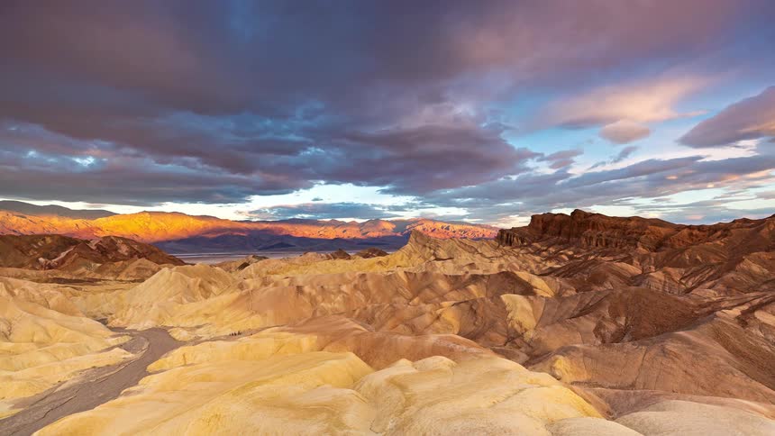 A view of a desert landscape shows many tan and brown rolling hills under a sky filled with dark purple and grey clouds that move quickly as the sun rises