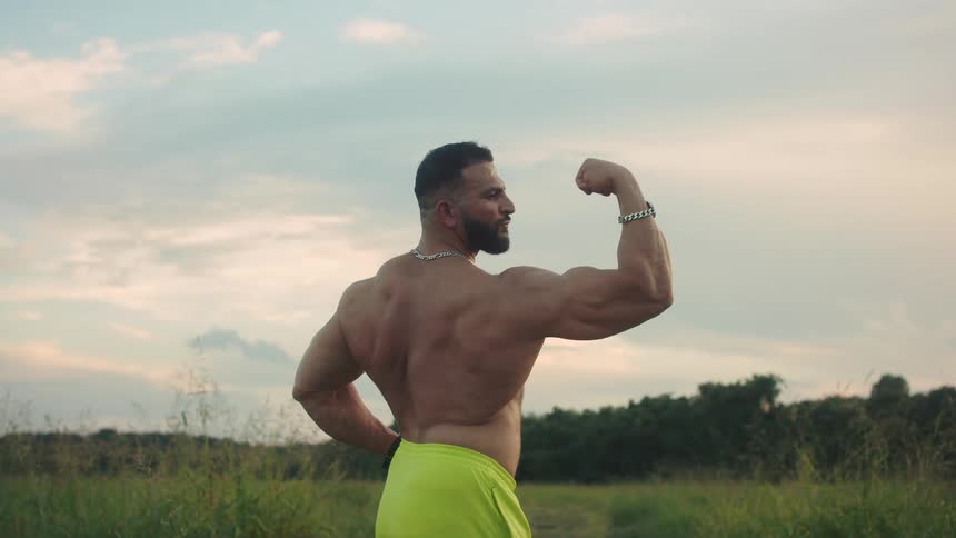 Muscular man seen from behind flexing his arm in an open area with vegetation in the background