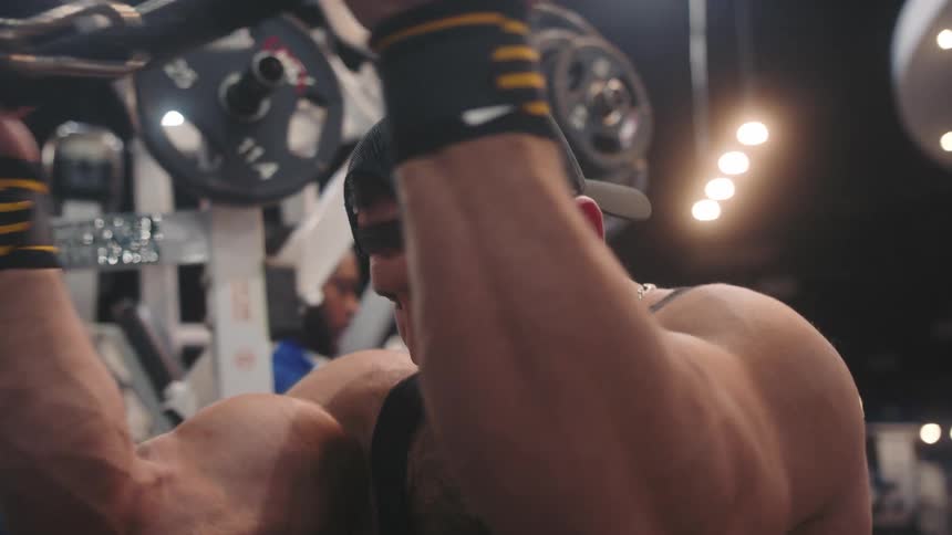 A close view of a muscular man with a beard as he performs bicep curls using a cable machine while sitting in a gym with various weight equipment