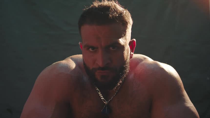 A close view of a muscular man with a beard and a silver chain necklace looking intensely toward the camera while sweat glistens on his skin in warm lighting