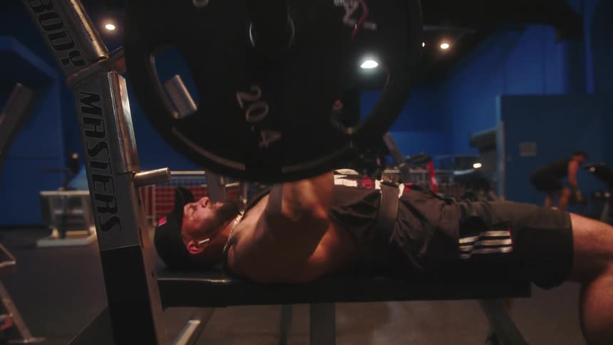 A muscular man with a beard wearing a black tank top and shorts lies on a weight bench and performs chest presses with a heavy barbell in a gym with blue walls and bright overhead lights