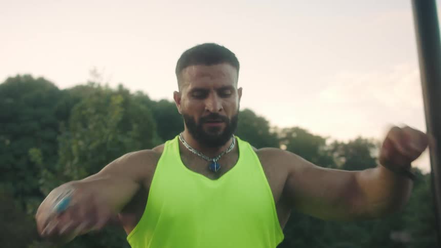 A muscular man with a beard wearing a neon yellow tank top and matching shorts performs stretches and joint rotations at an outdoor workout park with trees in the background