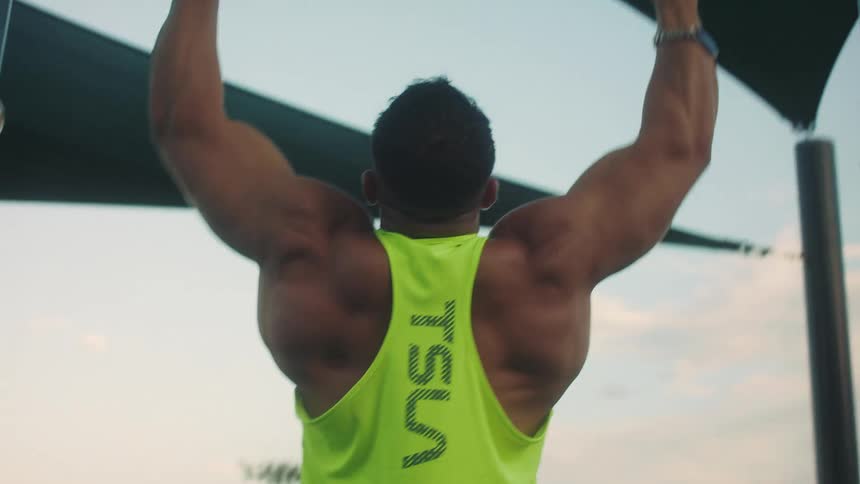 A view from the back shows a muscular man wearing a neon yellow tank top as he performs pullups on a metal bar at an outdoor gym