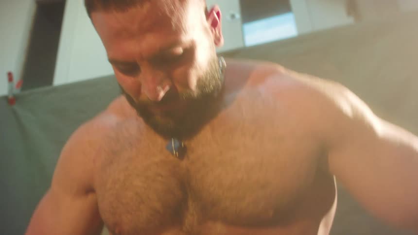 A muscular man with a beard and no shirt poses and flexes his muscles in a studio while wearing white shorts and a silver chain with a blue stone