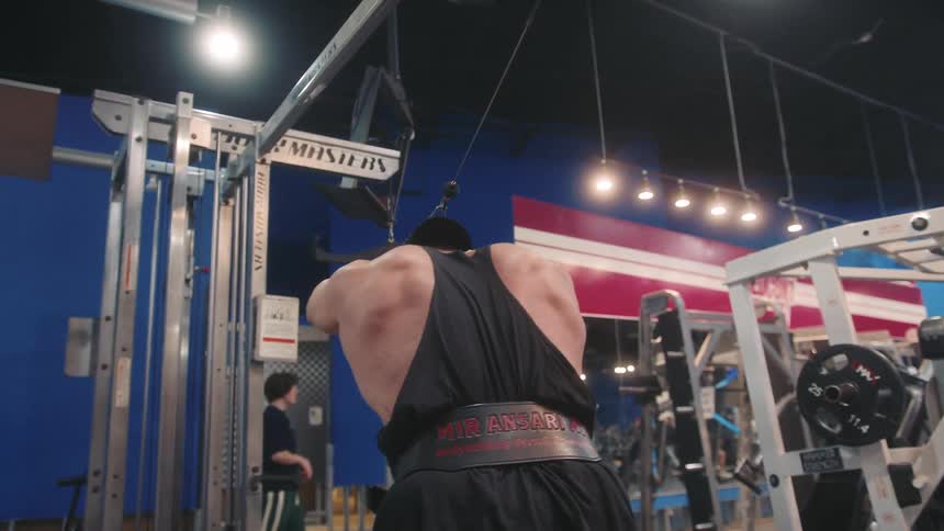 A muscular man wearing a lifting belt and wrist wraps performs overhead cable tricep extensions with a rope attachment inside a gym he faces away from the camera as he pulls the weight forward while standing in front of a cable machine station