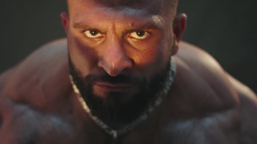 Muscular man faces forward in closeup with a beard metal necklace and dramatic lighting across the face
