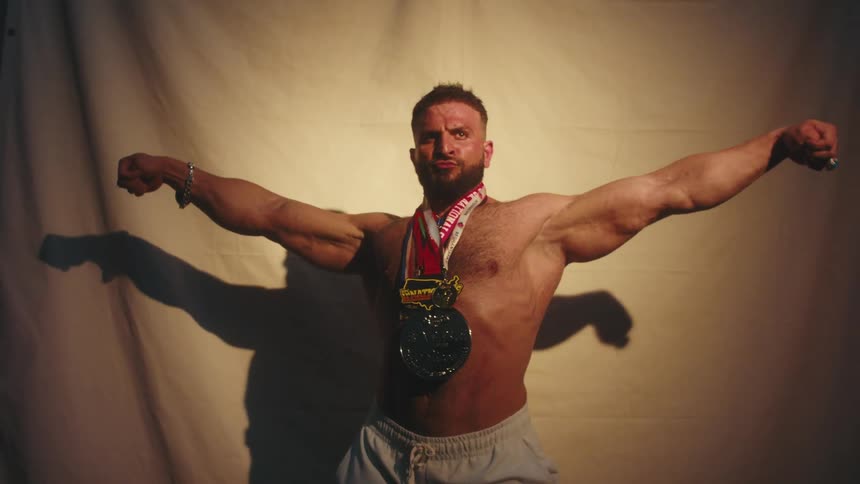 A close view of a muscular man with a beard and chest hair as he flexes his arms and torso while wearing several large medals around his neck in front of a plain fabric backdrop