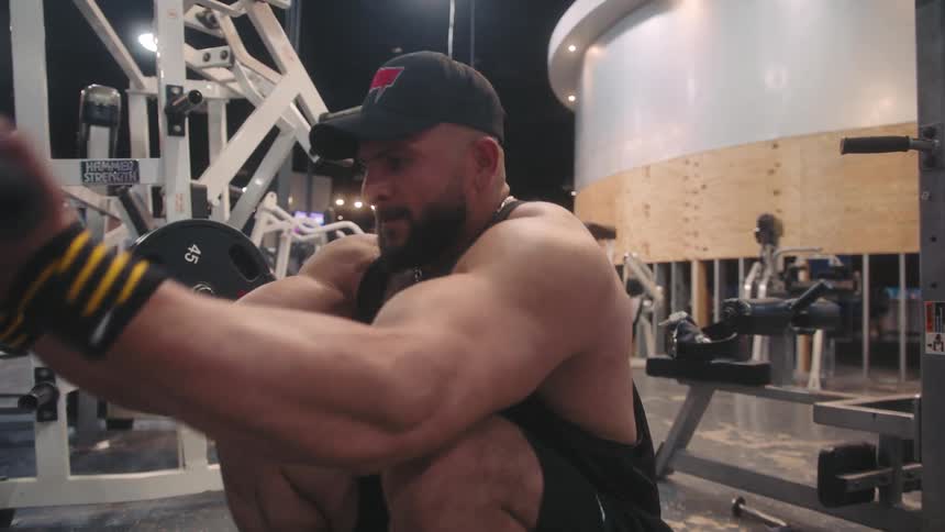 A muscular man wearing a black tank top and a baseball cap squats while performing bicep curls using a cable machine in a gym with various weight equipment