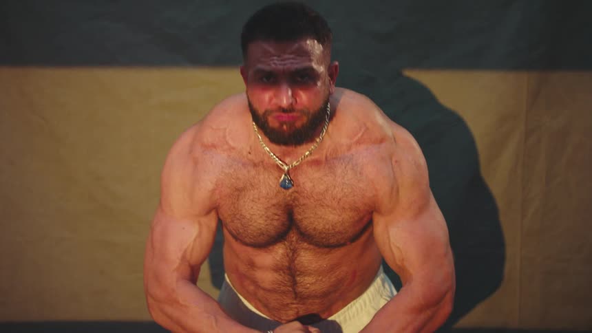 A muscular man wearing white shorts and a gold chain flexes his chest and arm muscles while posing against a dark backdrop in warm light