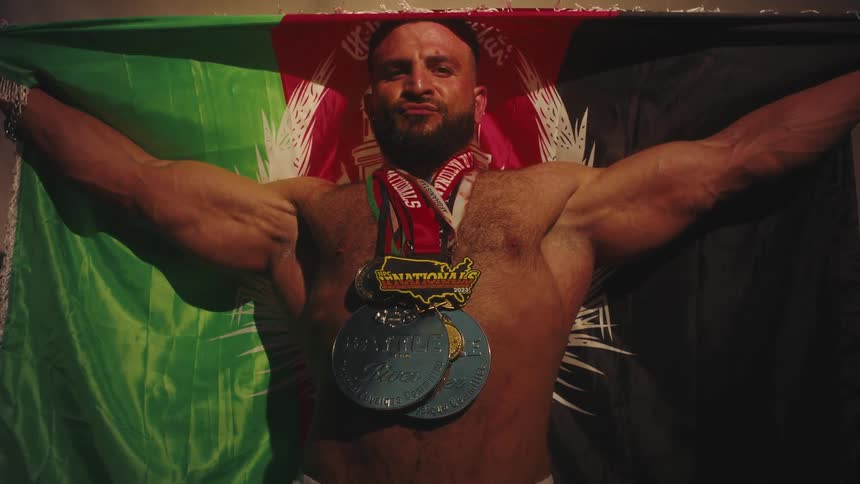 Muscular man displaying medals and a championship belt with a flag draped over his shoulders in an indoor setting