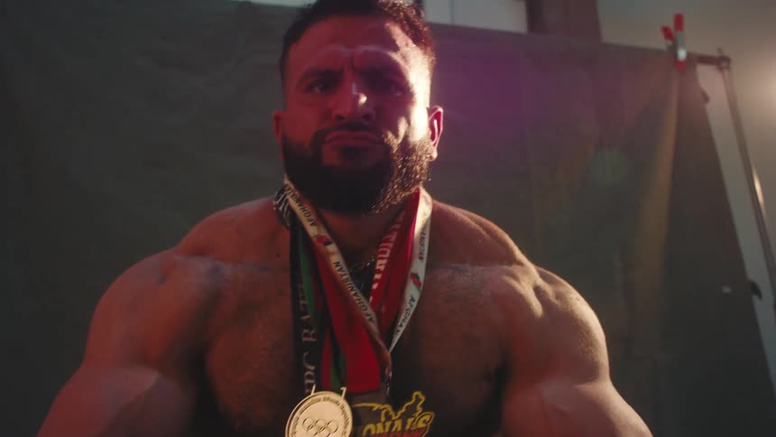 Bodybuilder seated with medals around his neck against a dark background