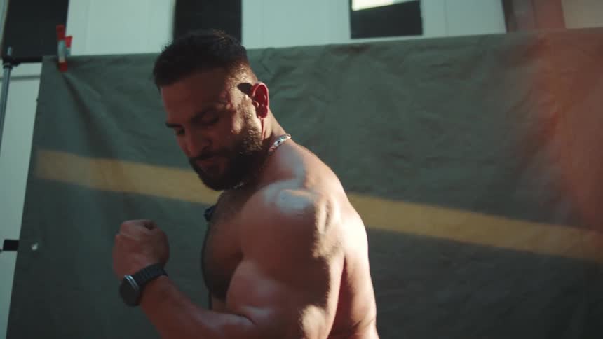 A bodybuilder flexes his huge muscles outdoors at midday shirtless in the bright sun