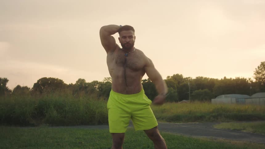 Muscular man posing with flexed arms in an open area at dusk