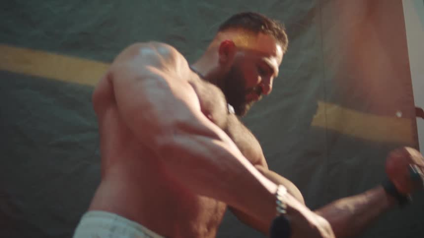 A muscular man with a beard and chest hair performs a bodybuilding posing routine in a studio