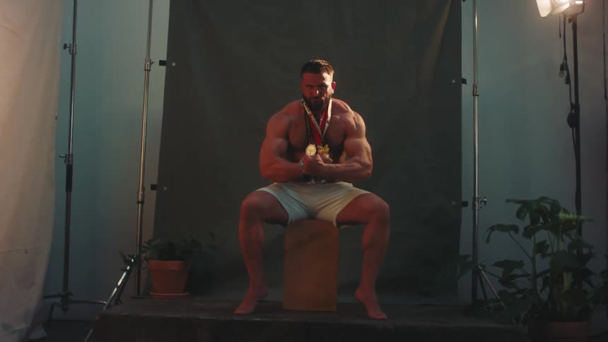 Muscular person poses seated with gold medals in a studio with a dark background and dramatic lighting