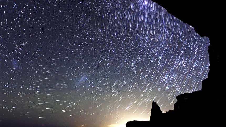 A time lapse view shows stars rotating in a circular motion across a dark night sky framed by the silhouette of a rocky cave opening