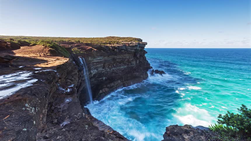 A waterfall drops from a rocky coastline at midday with the ocean water below turning a bright blue