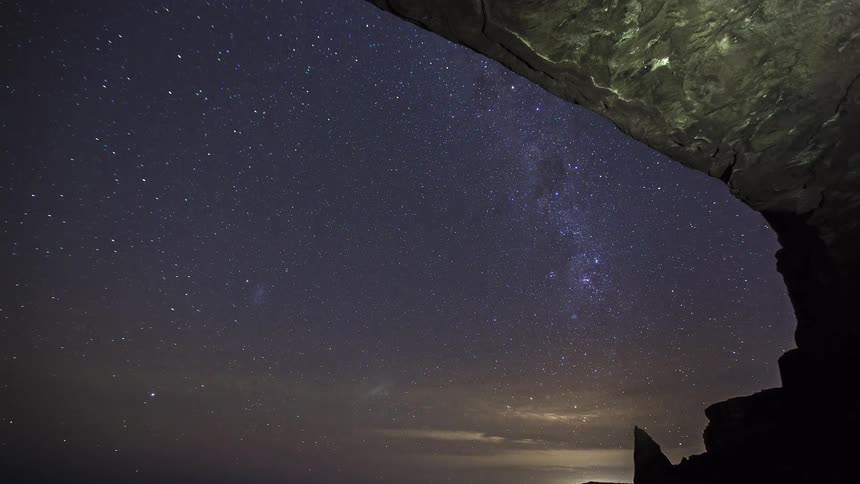 A view from the inside of a dark cave looks out at a vast night sky filled with many bright stars and the milky way galaxy as clouds drift along the horizon