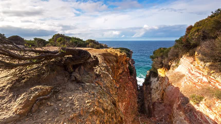 A view of a rocky coastline shows a deep narrow canyon leading to the ocean with layers of red and tan stone visible under a blue sky with white clouds