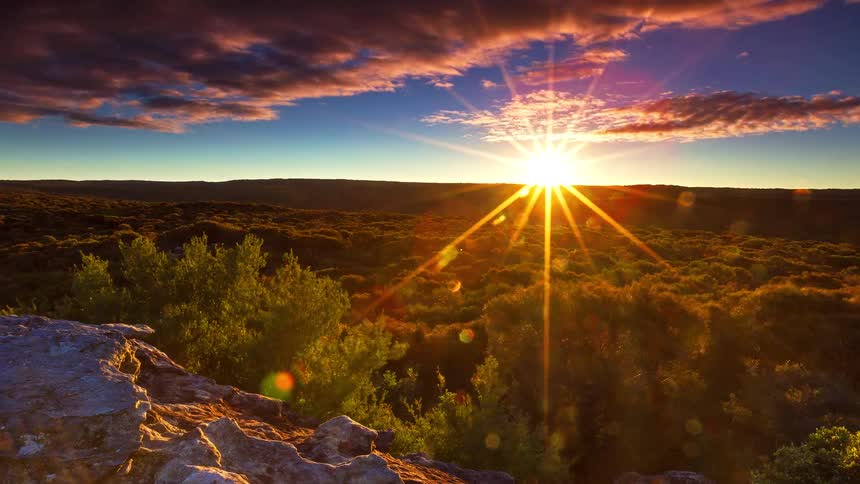 A view from a rocky ridge shows a wide landscape of green trees as the sun sets and the sky turns from blue to shades of orange and dark purple while clouds move quickly overhead