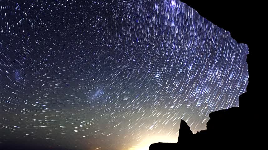 A view from inside a dark cave looks out at a vast night sky where many bright stars rotate and leave light trails as clouds and a glow appear on the horizon