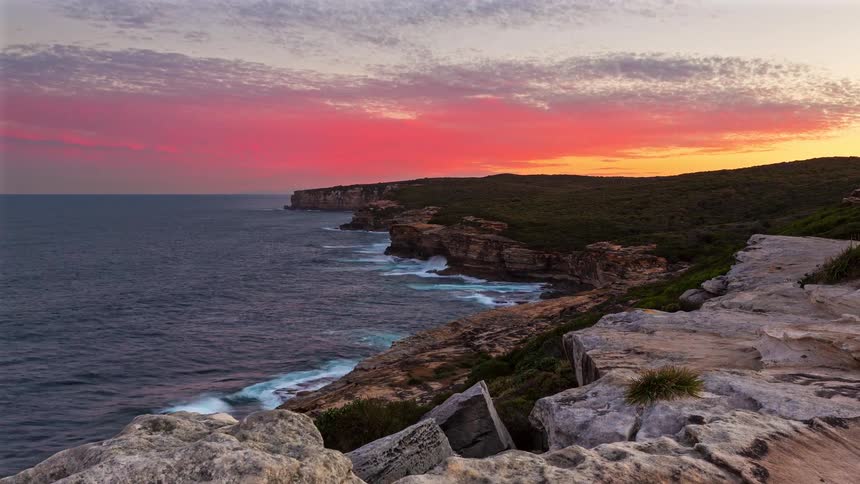 A dramatic sunset paints the sky in vibrant pinks and oranges over rugged coastal cliffs meeting the dark ocean waves