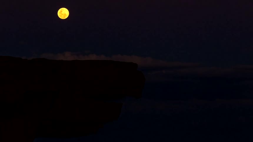 Full moon hangs above a silhouetted rock formation against a cloudy night sky