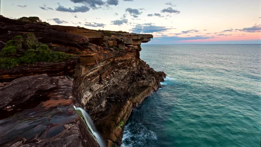 A view from above shows a rocky cliff over the ocean where a small waterfall flows into the water as the sun sets and a full moon rises into a purple and dark blue sky