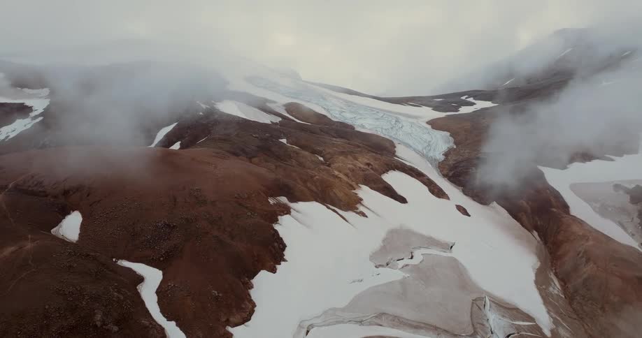 An aerial view shows a vast and dark mountain landscape with patches of white snow and a large glacier under thick grey fog